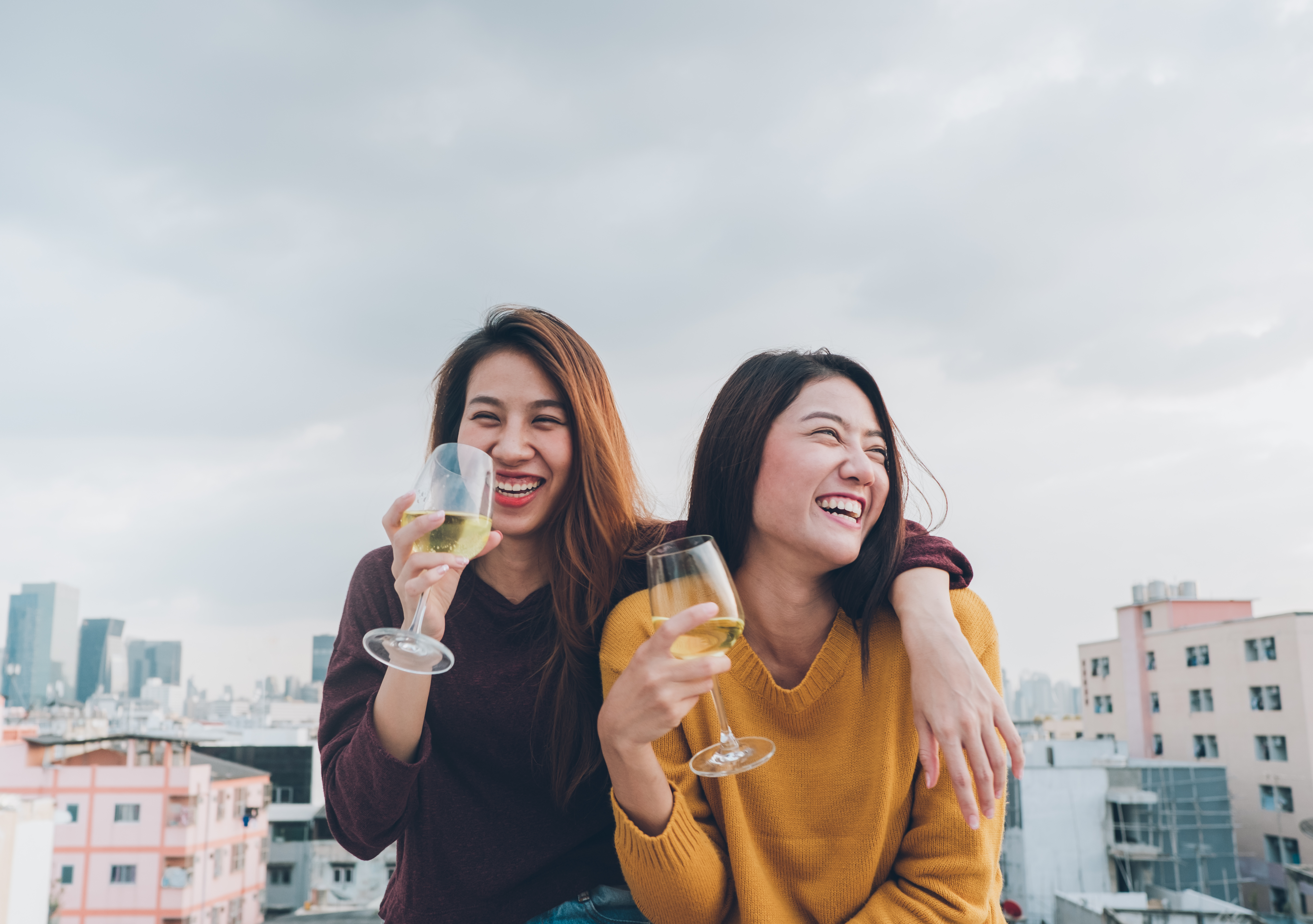 Two women on a casual date laughing on a rooftop holding glasses of white wine